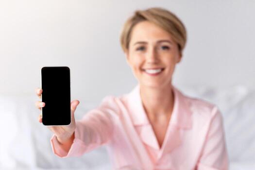 Closeup of smiling woman holding smartphone with black blank screen in hand, showing device, lady sitting on bed in blurred background. Selective focus on gadget with empty free space for mock up photo
