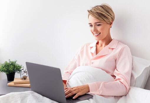 Portrait Of Smiling Beautiful Middle Aged Woman In Pink Sleepwear Sitting On Bed Under Blanket At Home, Using Computer Typing On Laptop Keyboard, Browsing Internet, Working Or Resting photo