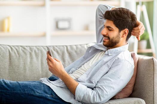 Leisure concept. Relaxed arab man using cellphone, browsing internet and texting lying comfortably on couch, side view. Smartphone user networking using social media application, free space photo