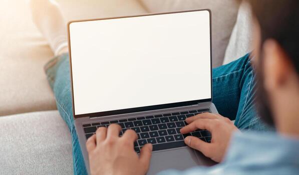 Technology concept. Unrecognizable guy using laptop computer with blank screen, surfing net and advertising internet website, sitting at home on sofa, over the shoulder view. Mockup, selective focus photo