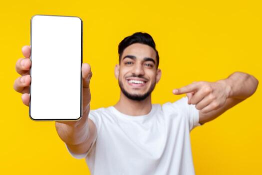 Happy bearded arab guy pointing at modern cellphone with empty screen in his hand, recommending newest mobile application or showing amazing offer over yellow studio background, mockup photo