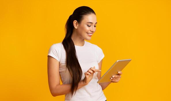 Charming Woman Using Digital Tablet And Browsing Internet Over Yellow Studio Background. Copy Space photo