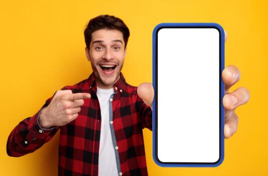 Recommendation. Portrait of excited guy holding big smartphone with white blank screen in hand, showing close to camera and pointing at device. Gadget with empty free space for mock up, yellow banner photo
