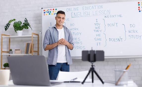 Easy English with tutor online during covid-19 pandemic. Millennial guy near blackboard with rules explain grammar, looks at web camera of smartphone on table with laptop in interior of class at home photo