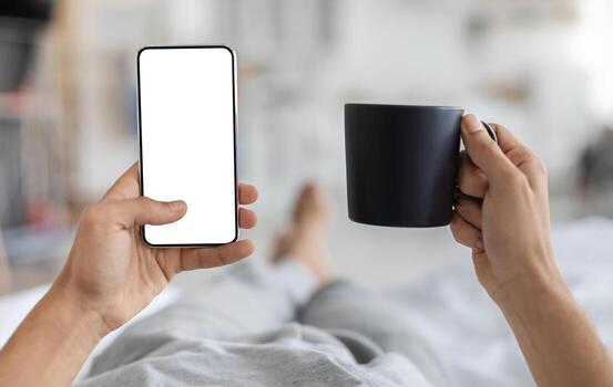 masculino manos participación teléfono inteligente con blanco pantalla y taza de café mientras acostado en cama a hogar, irreconocible hombre utilizando moderno artilugio con Copiar espacio para sitio web o móvil aplicación diseño, Bosquejo foto