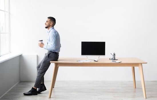 Smiling Successful Bearded Man In Formalwear Sitting Leaning On Table, Drinking Coffee Looking At Window, Showing Pc With Blank Black Mock Up Screen At Home Office, Free Copy Space, Full Body Length photo