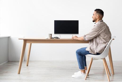 Happy Smiling Arab Man Sitting At Desk At Home Office Using Pc Computer With Blank Black Screen For Mock Up Template, Free Copy Space, Rear Back View. People, Technology, Remote Work Concept photo