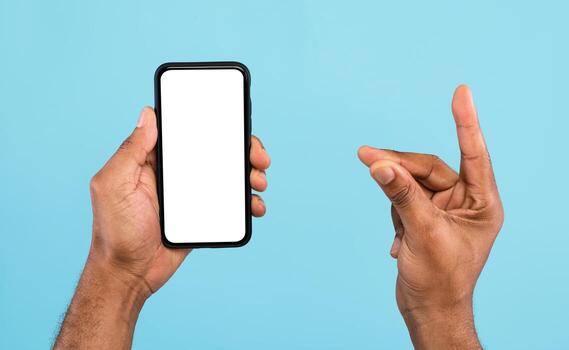Cropped view of young black guy showing smartphone with empty white screen and holding something on blue background, mockup for mobile app, website and business card design photo