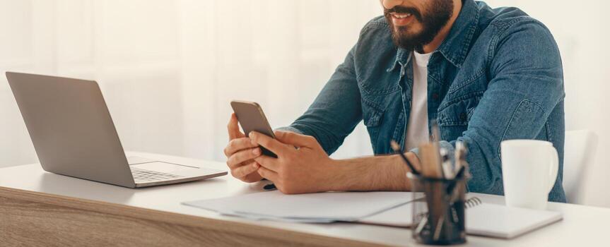Arab freelancer guy using cellphone while working on laptop at desk at home office, browsing new app or messaging with friends, sitting at workplace with computer, free space photo