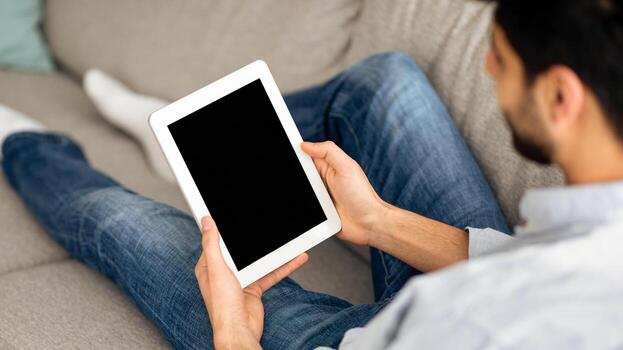 Advertisement concept. Millennial arab man using digital tablet with blank black screen, browsing website or showing new app, sitting on sofa at home. Mockup for your design photo