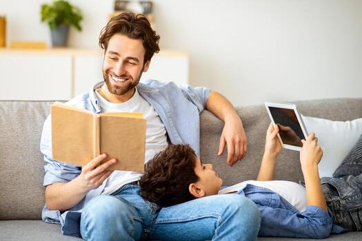 Different generations. Father reading book while his son using digital tablet, resting and sitting together on sofa. Leisure time, spending evening at home photo