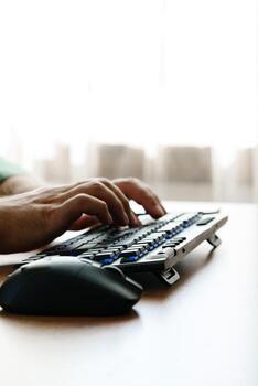 Hands typing on a computer keyboard while using a mouse near a window photo