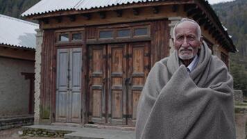 äldre man insvept i en värma filt står självsäkert i främre av en rustik trä- byggnad, visa upp elasticitet och karaktär i en lugn utomhus- miljö video