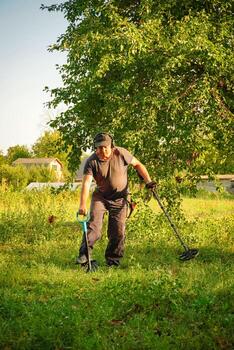 A man uses a metal detector in a green field, focused on finding hidden objects in the grass under the warm light of late afternoon. The area is surrounded by trees and distant structures photo
