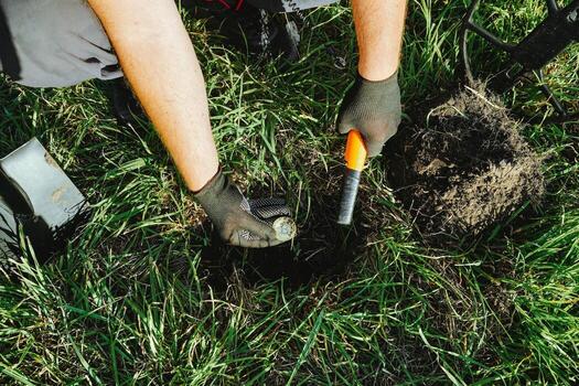Hands are seen working with a digging tool in grassy soil, focusing on uncovering possible items or artifacts. The scene is sunny and outdoor, showing a close-up of the activity photo