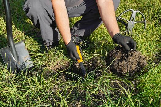 A person kneels in a vibrant green field, using a metal detector and digging into the soil. They are exploring for historical artifacts and valuable items photo