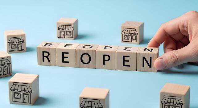 Hand arranging wooden blocks spelling REOPEN, surrounded by smaller blocks with shop icons, signifying business reopening and recovery on a light blue background photo