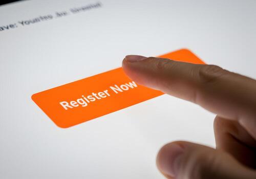 Closeup of a finger pressing an orange Register Now button on a touchscreen device signifying online registration digital interaction and immediate action photo