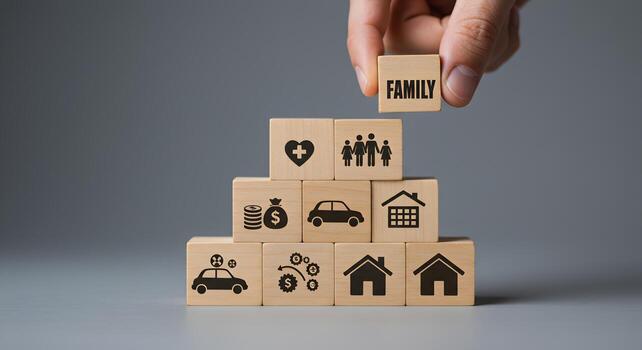 Hand placing wooden block with word FAMILY on top of a pyramid structure showing icons representing home car health and finances illustrating family values and building a secure future photo