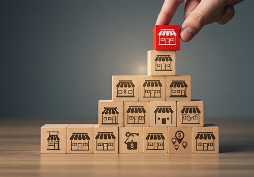 Hand placing a red block with store icon on top of a wooden block pyramid representing business growth and expansion strategy The concept illustrates increasing sales market share and building a succe photo