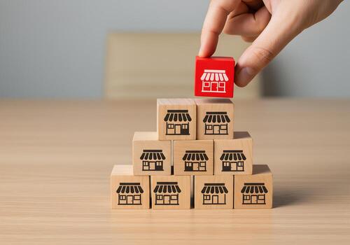 Hand placing red wooden block with store icon on top of a pyramid structure of similar blocks symbolizing business growth and expansion strategy This image represents concepts of retail ecommerce entr photo