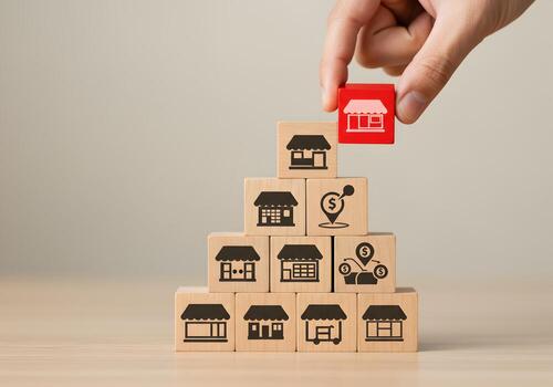 Hand placing red wooden block with store icon on top of a pyramid structure of wooden blocks with various store and location icons symbolizing business growth and expansion strategy photo