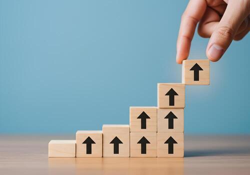 Hand placing wooden block with upward arrow on top of a growing stack of similar blocks on a table against a blue background symbolizing growth progress and upward trend photo