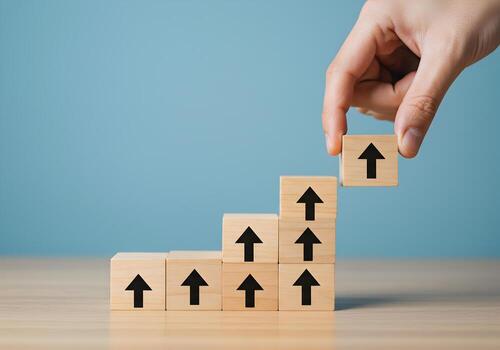 Hand placing wooden block with upward arrow on top of a growing stack of similar blocks on a table against a light blue background symbolizing growth progress and upward mobility photo
