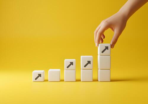Hand placing white cube with upward arrow on growing stack of similar cubes against yellow background symbolizing growth progress and success photo