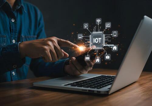 Person using smartphone and laptop to interact with IoT network showcasing smart home and device connectivity in a modern workspace setting The image conveys innovation technology and seamless integra photo
