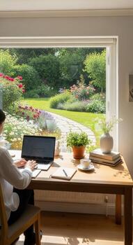 Woman working on a laptop at a desk with a view of a garden through a large window at daytime photo