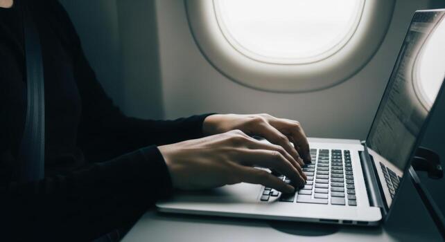 Person typing on a laptop computer while sitting on an airplane near a window during a flight photo