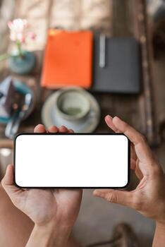 Hands holding a smartphone with a blank screen above a table with coffee and notebooks, ready for customization. photo