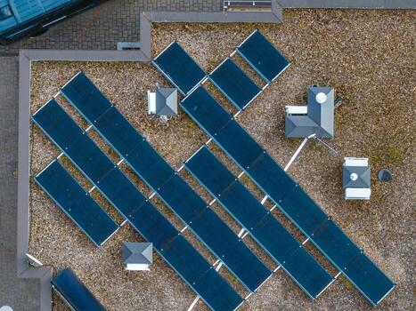 Aerial view of a rooftop with solar panels in a grid pattern, gravel surface, small structures, and a paved area with a parked vehicle visible. photo