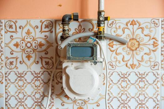 A gas meter with a digital display is mounted on a peach colored wall with intricate white and orange patterned tiles, connected to pipes and a hose. photo
