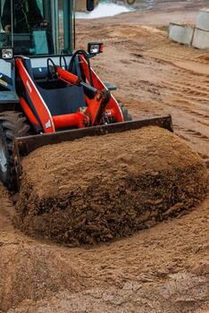 A red and white front loader operates on a dirt surface, its bucket filled with sand. Tire tracks and concrete blocks are visible in the industrial setting. photo