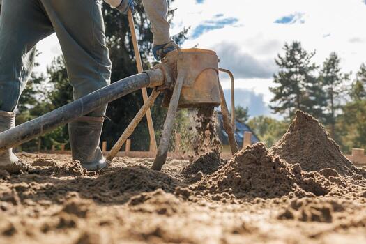A worker in protective gear operates a sandblasting machine, creating sand piles on a sandy surface. Trees are visible in the distance under clouds. photo