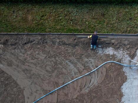A worker levels a sandy surface using a tool, with a long hose extending across the area. A strip of grass is visible at the top of the frame. photo