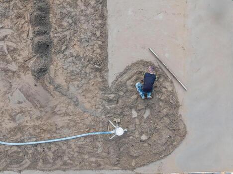 A worker kneels on a surface, spreading and leveling wet concrete with a long tool. A hose and tripod like device are visible near the work area. photo