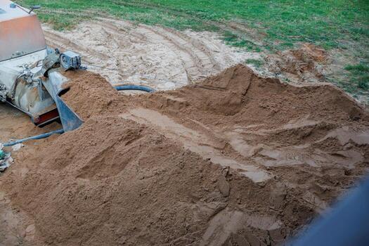 A large sand pile at a construction site with visible tire tracks, bordered by grass. Machinery with a hose is partially visible on the left side. photo