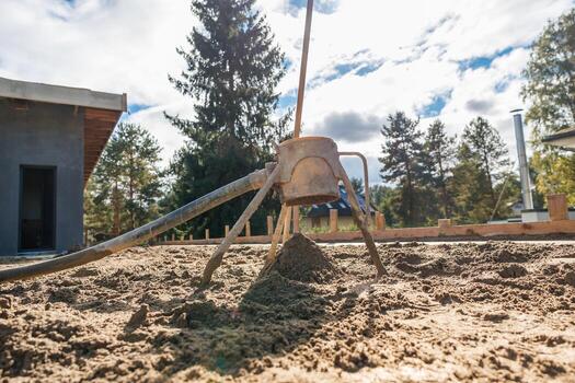 A concrete leveling tool is placed on sandy ground at a construction site with tall evergreen trees, a partially built structure, and a cloudy sky. photo