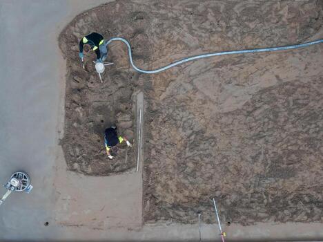 Two workers operate tools on a partially prepared surface at a construction site. Wet and dry soil, a leveling device, and poles are visible. photo