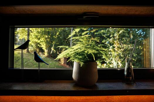 A windowsill displays a potted fern in a ribbed ceramic planter, bird figurines on stands, and a glass diffuser with reeds, with trees visible outside. photo