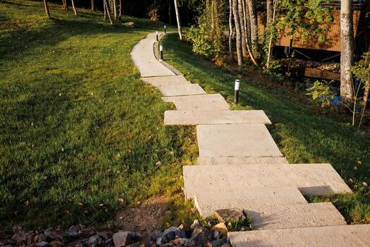 A concrete pathway with irregular slabs curves through grass bordered by trees. Modern light posts line the path, with a wooden structure to the right. photo