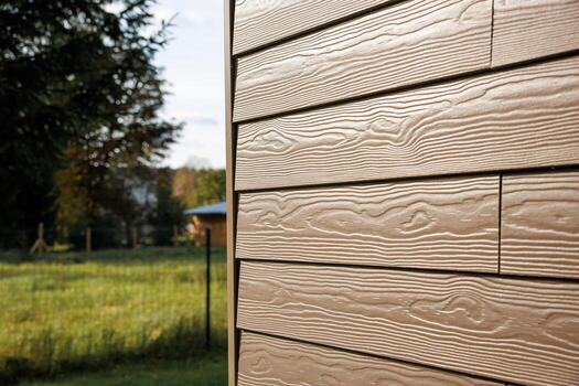 Textured beige horizontal siding with visible wood grain pattern, grassy area with fence, trees, and a small structure in the distance. photo