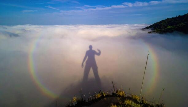 Cinematic mountain photo of a Brocken spectre projected onto valley fog from a ridge, a faint circular glory ring around a shadow for ethereal mood.