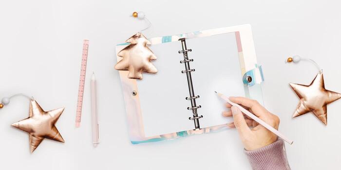 Female writing in copybook or diary with bright cover on white background, shiny Christmas tree and star. Concept of planning personal future goals and ideas for New Year, top view banner photo