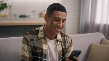 sonriente africano americano étnico hombre chico masculino mensajería con móvil teléfono a hogar disfrutando teléfono inteligente compras en línea teléfono aplicación participación hojeada comercio electrónico servicios golpe fuerte red web utilizando en Departamento video