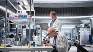 gammal man forskare använder sig av mikroskop med en kollega för klinisk studie. glidning de prov bricka för undersökning och stödjande genetik forskning, Avancerad analys för sjukvård. kamera a. video