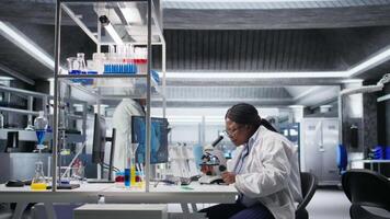 African American scientist focuses on a microscope with tray of samples in modern laboratory. Using observation tool for diagnostics, molecular analysis and discovery. Optics in bioscience. Camera A. video